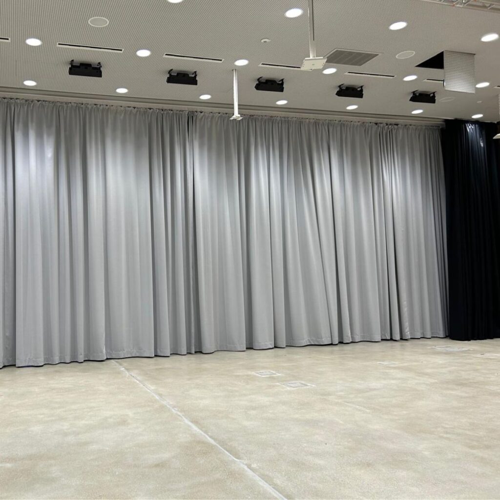 Kleine Bühne mit geschlossener, blauer Bühnenvorhang und professioneller Deckenbeleuchtung.