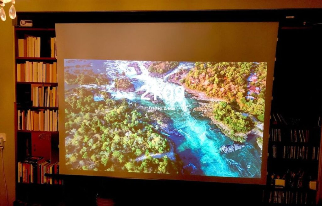 Projizierte Landschaft auf einer 300 cm Rückpro-Aufpro Motorleinwand mit schwarzem Gehäuse, front abrollend zur Regalmontage (Herstellung nach Spezialmaß) in einem privaten Wohnzimmer